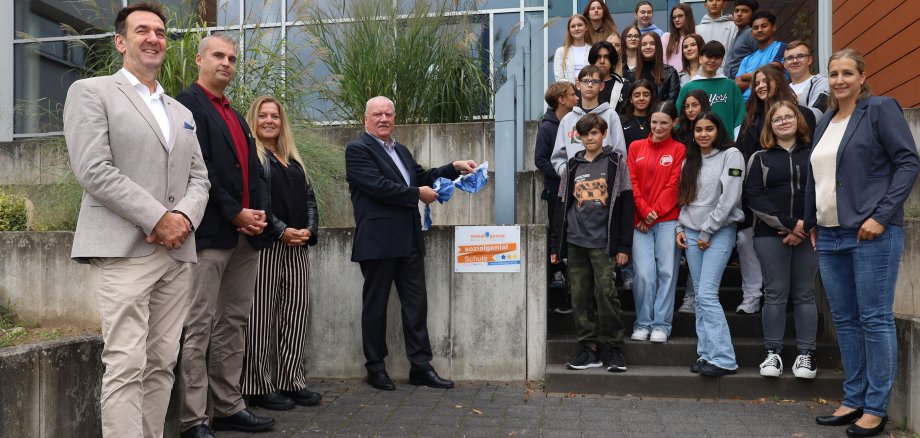 Auf dem Foto von Links: Schulleiter Hauke Wagner, Fördervereinsvorsitzender Michael Heng, Schulelternbeirätin Claudia Klein sowie Frank Lortz (Vizepräsident des Hessischen Landtags) bei der Enthüllung der Auszeichnung zur sozialgenial Schule mit der Lehrkraft Frau Smith und der projektbeteiligten Schülerschaft.